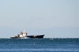 A large cargo ship sailing on a slightly choppy sea, with a distant mountain range visible under a clear sky. The ship appears to be moving steadily across the water, generating small waves along its sides.