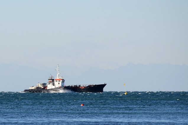 A large cargo ship sailing on a slightly choppy sea, with a distant mountain range visible under a clear sky. The ship appears to be moving steadily across the water, generating small waves along its sides.