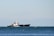 A large cargo ship sailing on a slightly choppy sea, with a distant mountain range visible under a clear sky. The ship appears to be moving steadily across the water, generating small waves along its sides.