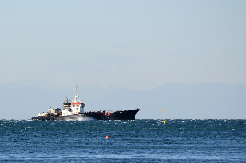 A large cargo ship sailing on a slightly choppy sea, with a distant mountain range visible under a clear sky. The ship appears to be moving steadily across the water, generating small waves along its sides.
