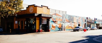 A street scene featuring a building painted in yellow and black with a sign for a smoke shop. Adjacent to it is a large mural with geometric patterns and vibrant colors depicting various abstract shapes. Several cars are parked along the street, and a person in a red coat walks across the road. The scene is illuminated by natural daylight, with a tree visible on the left side of the image.