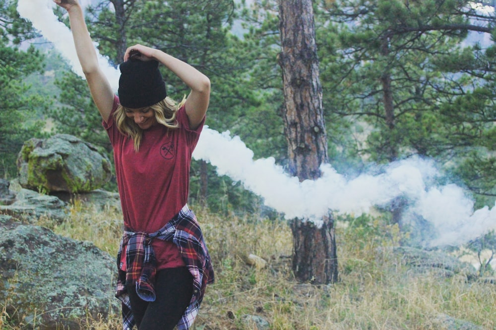 Frau hebt Arme mit weißer Rauchbombe, die tagsüber neben einem von Bäumen umgebenen Felsen steht