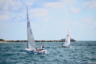 Two small sailboats with single sailors navigate the open sea. The sky is partly cloudy, and there is a stone breakwater in the background. The water is a deep blue with gentle waves, suggesting a calm day for sailing.