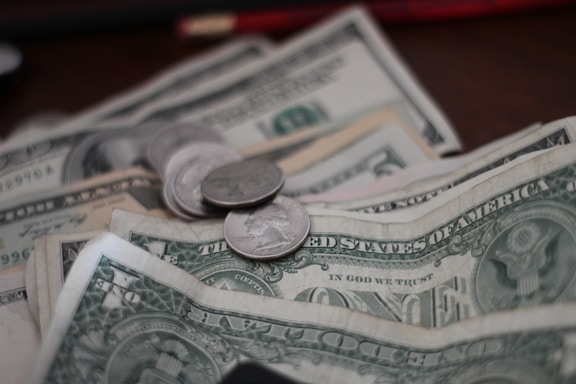 A close-up view of several US dollar bills and coins arranged on a surface. The image prominently features one-dollar bills and several quarters. The faded background indicates some currency notes are stacked slightly out of focus.