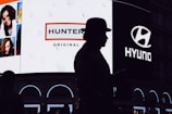 A silhouette of a person wearing a hat stands in front of a brightly lit billboard featuring advertisements for Hunter and Hyundai. The billboard also displays several portrait images of people.