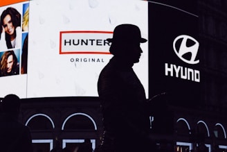 A silhouette of a person wearing a hat stands in front of a brightly lit billboard featuring advertisements for Hunter and Hyundai. The billboard also displays several portrait images of people.