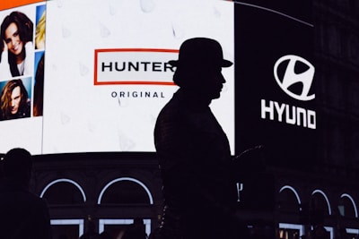 A silhouette of a person wearing a hat stands in front of a brightly lit billboard featuring advertisements for Hunter and Hyundai. The billboard also displays several portrait images of people.