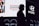 A silhouette of a person wearing a hat stands in front of a brightly lit billboard featuring advertisements for Hunter and Hyundai. The billboard also displays several portrait images of people.