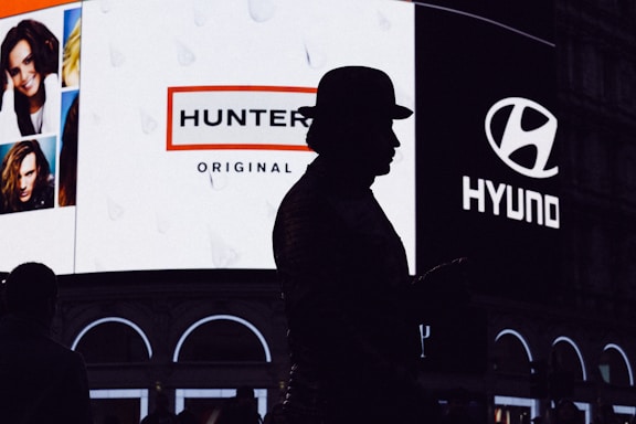 A silhouette of a person wearing a hat stands in front of a brightly lit billboard featuring advertisements for Hunter and Hyundai. The billboard also displays several portrait images of people.