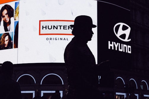 A silhouette of a person wearing a hat stands in front of a brightly lit billboard featuring advertisements for Hunter and Hyundai. The billboard also displays several portrait images of people.