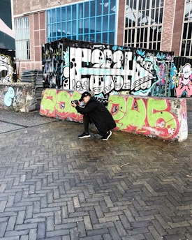 A person is crouching on a paved area while taking a photograph. The setting features vibrant graffiti art on a series of barriers and metal containers. The background includes a red brick building with large windows.