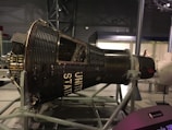 A space capsule displayed in a museum setting, supported by a metal framework. The capsule is labeled with 'UNITED STATES' and has a vintage, industrial design featuring numerous rivets and panels. The museum environment includes soft lighting and a visitor is visible in the background.