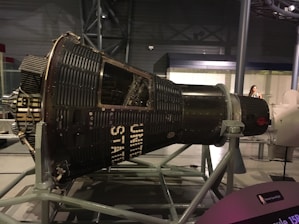 A space capsule displayed in a museum setting, supported by a metal framework. The capsule is labeled with 'UNITED STATES' and has a vintage, industrial design featuring numerous rivets and panels. The museum environment includes soft lighting and a visitor is visible in the background.