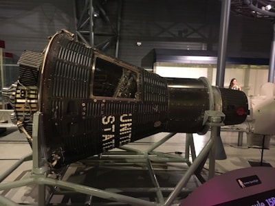 A space capsule displayed in a museum setting, supported by a metal framework. The capsule is labeled with 'UNITED STATES' and has a vintage, industrial design featuring numerous rivets and panels. The museum environment includes soft lighting and a visitor is visible in the background.