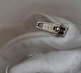 A close-up of a metallic zipper pull with embossed characters, positioned on a white fabric, possibly part of a garment or bag.