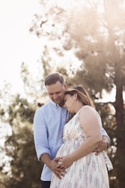 A couple stands closely together in a natural outdoor setting, with the woman gently cradling her pregnant belly. The man, wearing a light blue shirt, tenderly embraces her from behind. Sunlight filters through the trees, creating a warm, serene atmosphere.