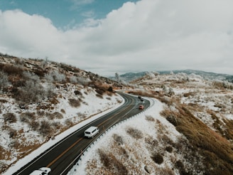 A winding road cuts through a snowy, mountainous landscape. Cars are traveling along the road, surrounded by hills with a light dusting of snow. The sky is overcast with clouds, and the scenery is marked by a mix of white snow and brown vegetation.
