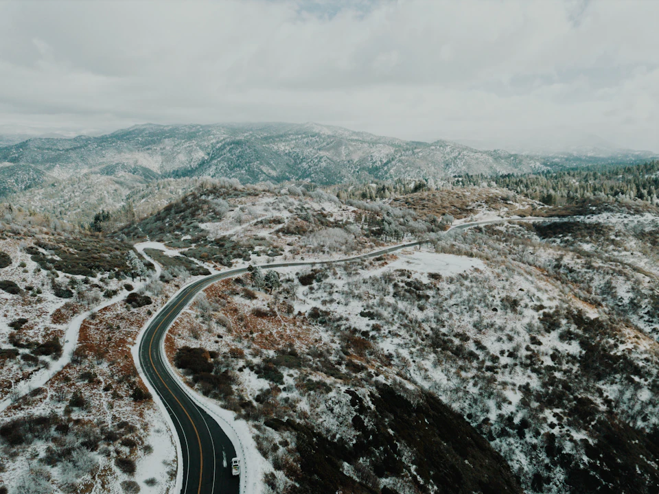 The image presents an aerial view of a winding road cutting through a mountainous landscape. The road, which is black and curving, meanders through the mountainside, disappearing into the distance. The surrounding area is covered in snow, with patches of grass visible on either side of the road. The sky above is overcast, casting a soft light over the scene.
