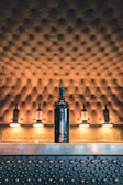 A bottle of wine is placed on a metallic counter adorned with raised circular patterns. The background features a tufted, upholstered surface with warm lighting, giving a luxurious and elegant atmosphere.
