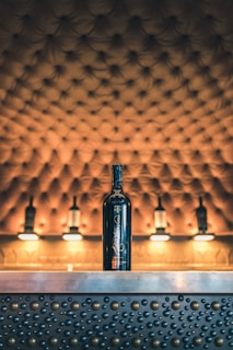 A bottle of wine is placed on a metallic counter adorned with raised circular patterns. The background features a tufted, upholstered surface with warm lighting, giving a luxurious and elegant atmosphere.