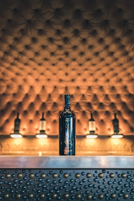 A bottle of wine is placed on a metallic counter adorned with raised circular patterns. The background features a tufted, upholstered surface with warm lighting, giving a luxurious and elegant atmosphere.