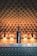 A bottle of wine is placed on a metallic counter adorned with raised circular patterns. The background features a tufted, upholstered surface with warm lighting, giving a luxurious and elegant atmosphere.