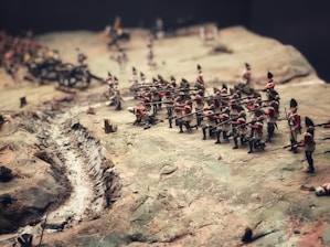 A diorama depicting a historic battle scene with miniature soldiers in red uniforms carrying rifles. The terrain is uneven, featuring a narrow trench or path along the battlefield and various small terrain elements.