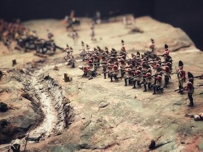 A diorama depicting a historic battle scene with miniature soldiers in red uniforms carrying rifles. The terrain is uneven, featuring a narrow trench or path along the battlefield and various small terrain elements.