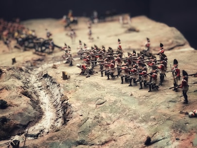 A diorama depicting a historic battle scene with miniature soldiers in red uniforms carrying rifles. The terrain is uneven, featuring a narrow trench or path along the battlefield and various small terrain elements.