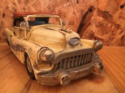 A vintage, worn-out toy car with a weathered yellow exterior sits on a wooden surface. The car features a prominent front grille and round headlights. The background consists of a wooden texture that complements the rustic look of the toy car.