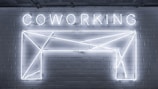 Neon lights form the word 'COWORKING' and an abstract geometric shape mounted on a brick wall. The design is sleek and modern, with a minimalistic style.
