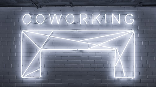 Neon lights form the word 'COWORKING' and an abstract geometric shape mounted on a brick wall. The design is sleek and modern, with a minimalistic style.