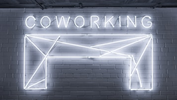 Neon lights form the word 'COWORKING' and an abstract geometric shape mounted on a brick wall. The design is sleek and modern, with a minimalistic style.