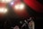 A musician is performing on stage with a guitar, surrounded by red drapes and illuminated by bright chandeliers. The scene includes other people, possibly band members or audience, with one person adjusting glasses.