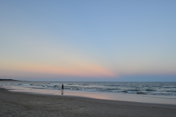 A serene beach scene with a lone person leisurely fishing by the shoreline under a vast, colorful evening sky. Soft hues of pink and blue blend as dusk approaches, creating a tranquil atmosphere. Gentle waves roll onto the sandy shore as the silhouette of the fisher lines the water's edge.
