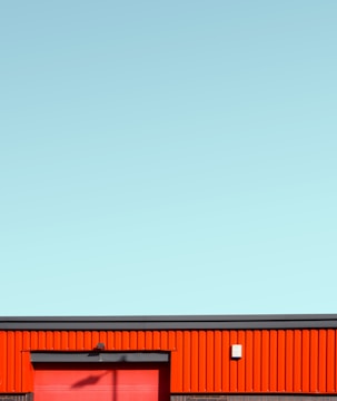 A minimalistic building facade featuring a bold red panel wall with a matching red door, set against a clear, pale blue sky. The structure is geometric, with horizontal lines and a few small fixtures visible.
