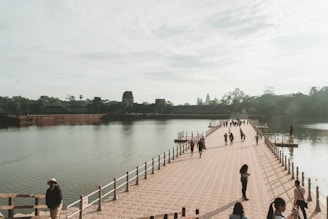 Pilgrims crossing a wooden bridge over a flowing river, framed by lush greenery and temple silhouettes.