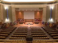 A large, formal conference room with numerous seats and desks organized in rows facing a central stage. The stage area is adorned with a large projection screen, and several microphones and symbols are present. Numerous international flags line the walls on either side of the stage. The room features warm lighting and a minimalist design with emphasis on functionality.
