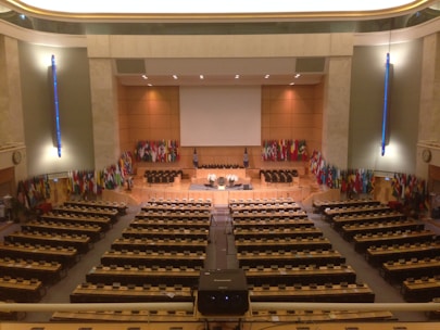 A large, formal conference room with numerous seats and desks organized in rows facing a central stage. The stage area is adorned with a large projection screen, and several microphones and symbols are present. Numerous international flags line the walls on either side of the stage. The room features warm lighting and a minimalist design with emphasis on functionality.