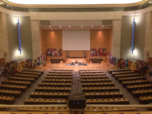 A large, formal conference room with numerous seats and desks organized in rows facing a central stage. The stage area is adorned with a large projection screen, and several microphones and symbols are present. Numerous international flags line the walls on either side of the stage. The room features warm lighting and a minimalist design with emphasis on functionality.