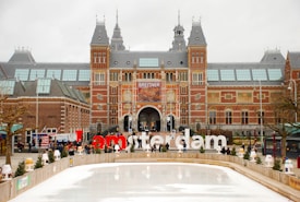 An expansive historic building with a red brick facade and intricate architectural details, featuring a large arched entrance. In front of the building is an ice skating rink, and above it, large letters spell out the word 'Iamsterdam' in a bold red and white font. People are seen walking around and gathering near the area.