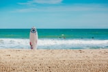 A sunny beach with a white surfboard sticking upright in the sand. The ocean is in the background with gentle waves and several people swimming or surfing. The sky is clear with some light clouds.