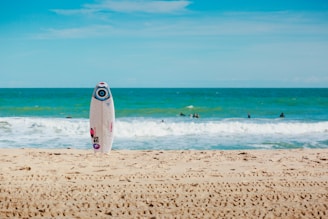 A sunny beach with a white surfboard sticking upright in the sand. The ocean is in the background with gentle waves and several people swimming or surfing. The sky is clear with some light clouds.