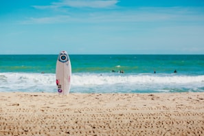 A sunny beach with a white surfboard sticking upright in the sand. The ocean is in the background with gentle waves and several people swimming or surfing. The sky is clear with some light clouds.