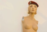 A mannequin upper torso with a stylized, artistic appearance, wearing a red beret tilted slightly to one side. The mannequin appears to be missing arms, with visible screws where they would attach. It has short, blonde hair styled in a blunt cut.