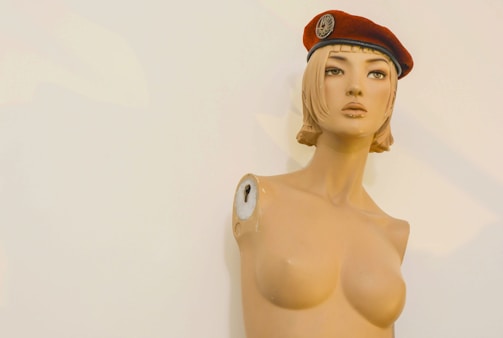 A mannequin upper torso with a stylized, artistic appearance, wearing a red beret tilted slightly to one side. The mannequin appears to be missing arms, with visible screws where they would attach. It has short, blonde hair styled in a blunt cut.