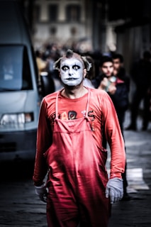 A person is dressed as a clown with black and white face paint and small tufts of hair on either side of the head. They are wearing a bright red jumpsuit and white gloves, standing in an urban setting with blurred people and vehicles in the background.