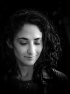 A black and white portrait of a person with long, curly hair, eyes gently closed, and a subtle smile. The lighting is soft, highlighting facial features and casting a serene expression.