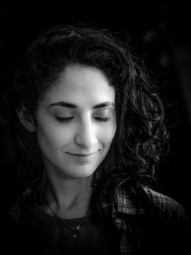 A black and white portrait of a person with long, curly hair, eyes gently closed, and a subtle smile. The lighting is soft, highlighting facial features and casting a serene expression.