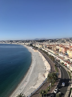A coastal cityscape with a long, curving beach bordered by clear blue water. A row of colorful buildings lines the road next to the beach, and a promenade with palm trees and people walking is visible. The area is bustling with activity, with a few parked cars and some people relaxing on the beach.
